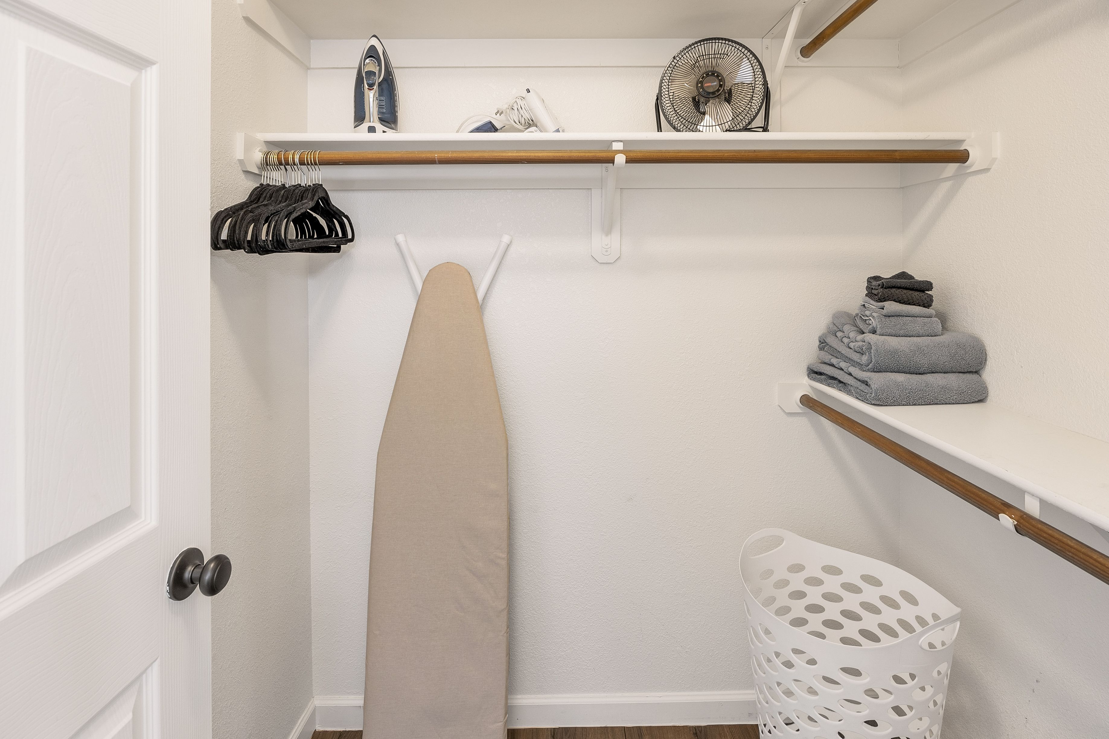 A laundry area with an ironing board, a fan, and a shelf with folded towels.