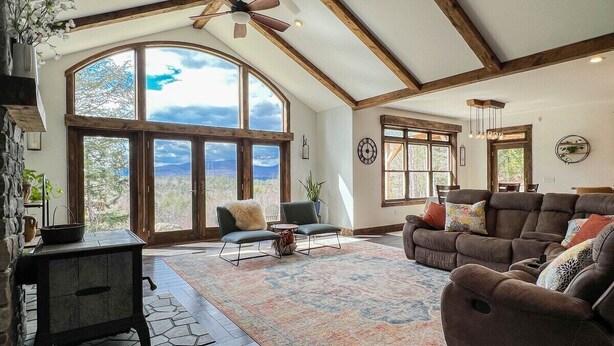 A spacious living room with a large window, a fireplace, and a ceiling fan. - R&R White Mountain Retreat, Hillside with Extraordinary Views! (Fryeburg)