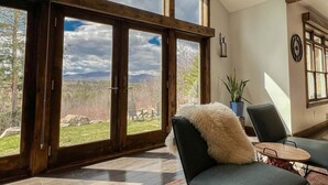 A room with large windows, a wooden ceiling, a chair with a fluffy pillow, and a small table. - R&R White Mountain Retreat, Hillside with Extraordinary Views! (Fryeburg)