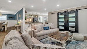 Un salon moderne avec un canapé, une table basse et un pouf. On y trouve également un coin repas avec une table et des chaises. La pièce est dotée d’une grande porte coulissante et d’une cuisine à l’arrière-plan.