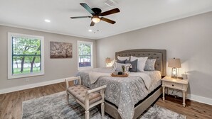 Une chambre avec un grand lit, un ventilateur de plafond, une table de chevet avec une lampe et une vue sur les arbres par la fenêtre.