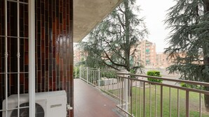 A balcony with a metal railing, a white air conditioning unit, and a view of trees and buildings.