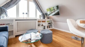 A modern living room with a glass coffee table, a sofa, a chair, a TV mounted on the wall, and a window with curtains. - Modern. Ruhig. Wiesenblick. - Ringhoog Hüs Whg. 5 "weitblick" - Objekt-id: Rhh/05 - Ringhoog Hüs (Sylt)