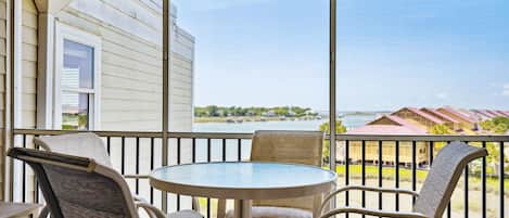 A covered outdoor area with a round table and four chairs, overlooking a body of water and buildings in the distance.