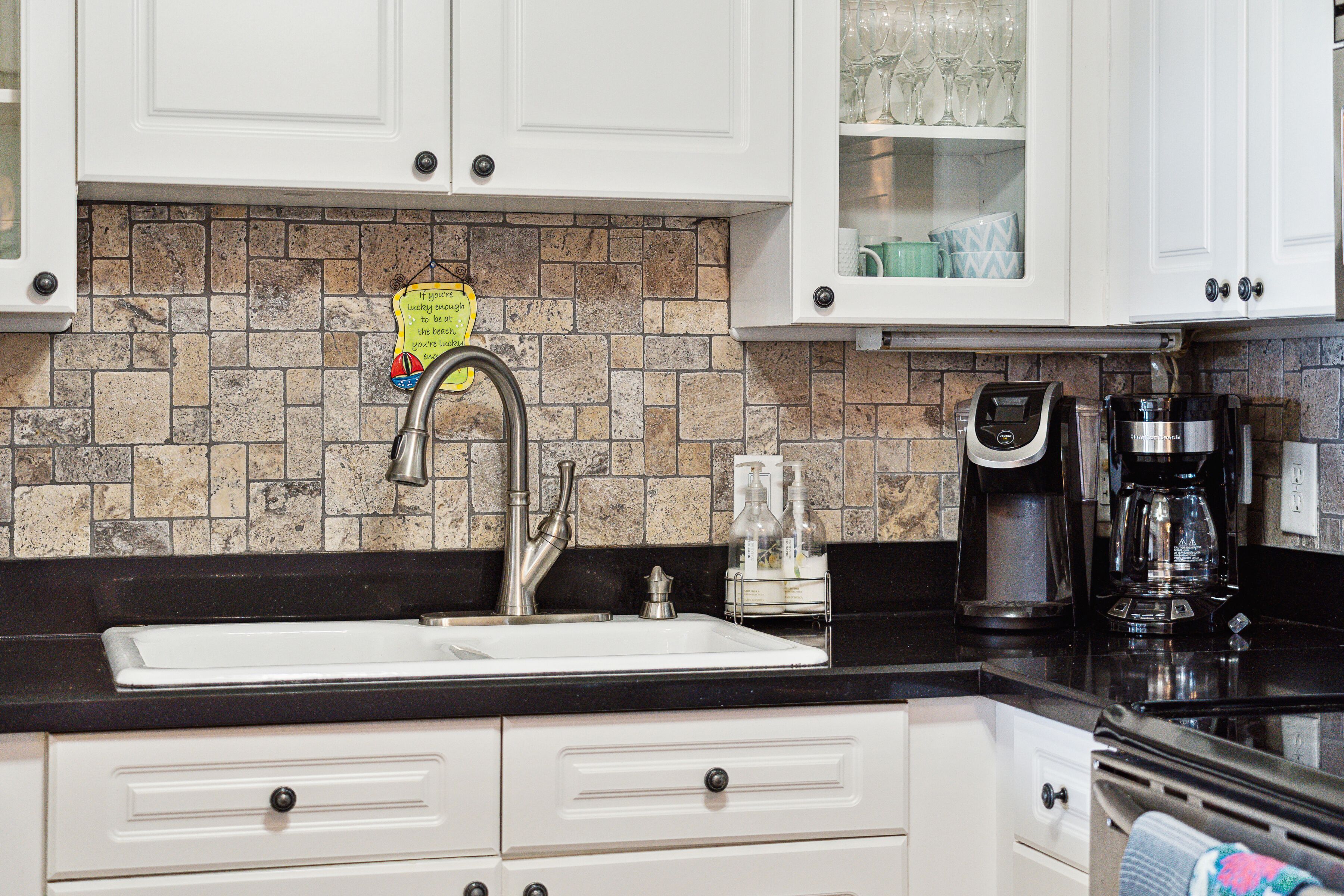 A kitchen with white cabinets, a black countertop, a stainless steel sink, a coffee maker, and a backsplash with patterned tiles.