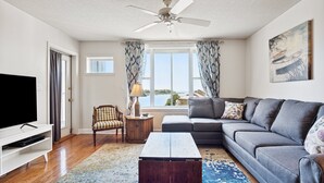 A living room with a grey sofa, a wooden coffee table, a TV on a white stand, a ceiling fan, and a large window with curtains.