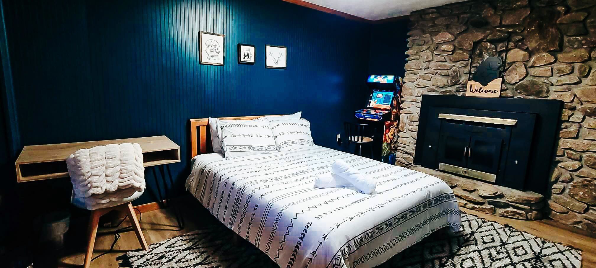 A bedroom with a bed, a small table with a towel, a stone fireplace, and framed pictures on the wall.