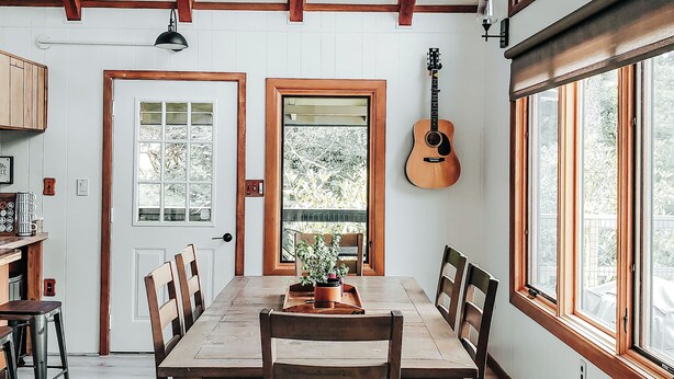 Uma sala de jantar com mesa de madeira, cadeiras e um candelabro. Há janelas e um violão na parede.