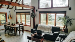 Uma sala de estar espaçosa com uma mesa de jantar de madeira, um sofá de couro preto e uma mesa de centro. Grandes janelas proporcionam luz natural e vista para o exterior.