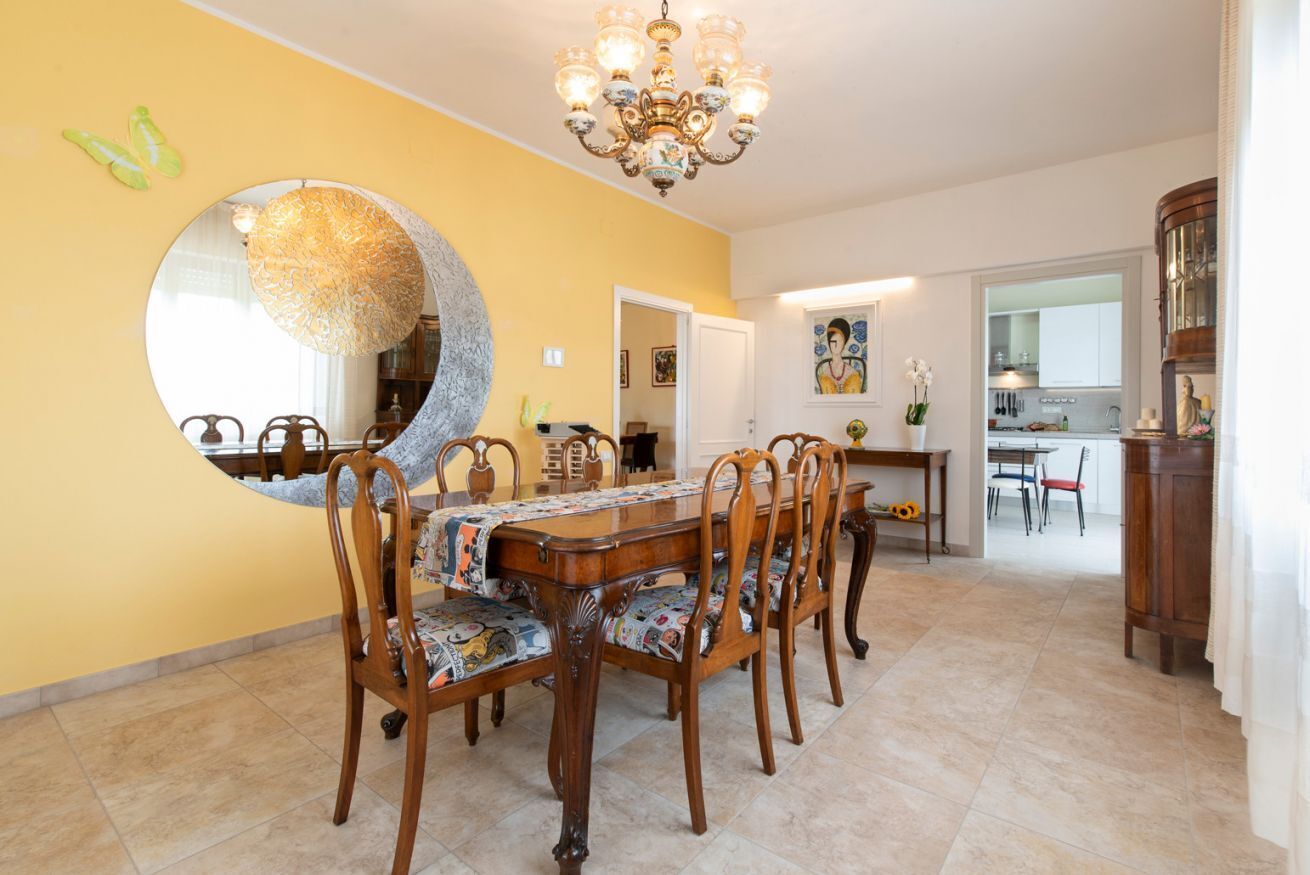 Una sala da pranzo con uno specchio rotondo, sedie di legno, un lampadario e una cucina sullo sfondo.