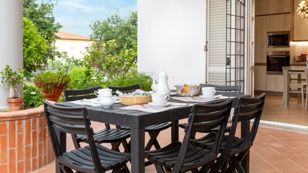 Un área de comedor al aire libre con una mesa preparada para cuatro personas, rodeada de sillas, con vistas a un jardín y un edificio al fondo.