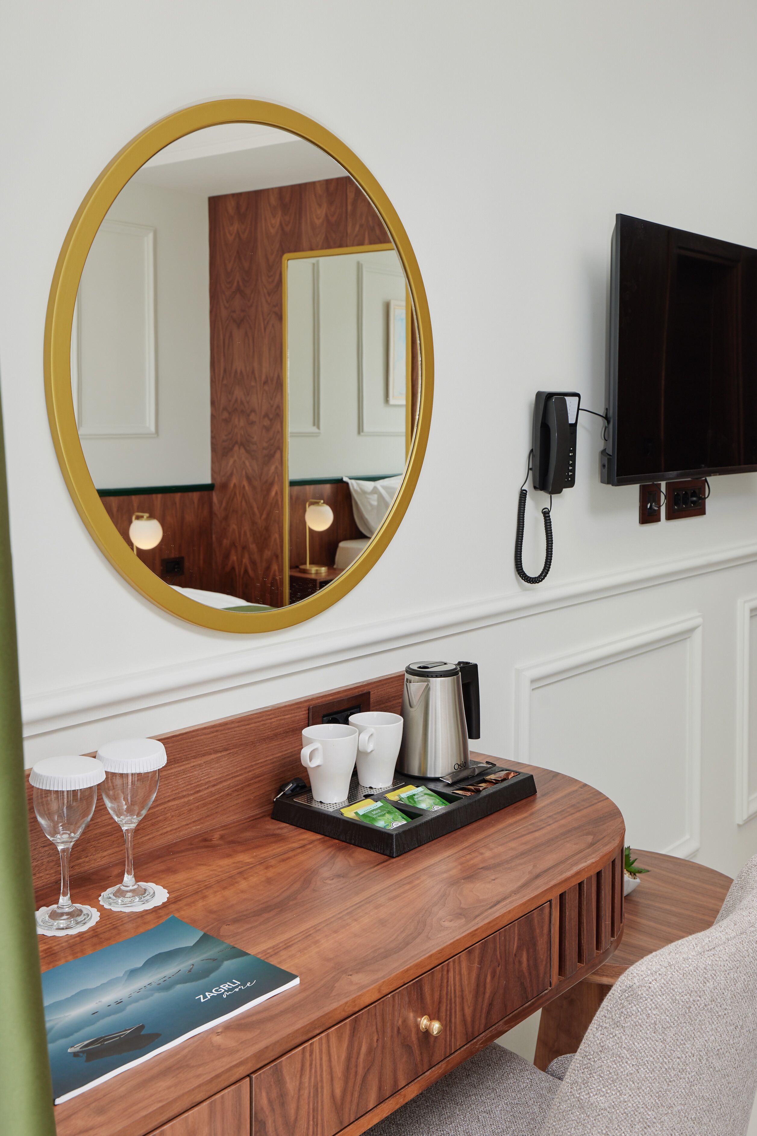 A hotel room with a wooden console table, a large mirror, a flat-screen TV, a telephone, and a tray with coffee and snacks.