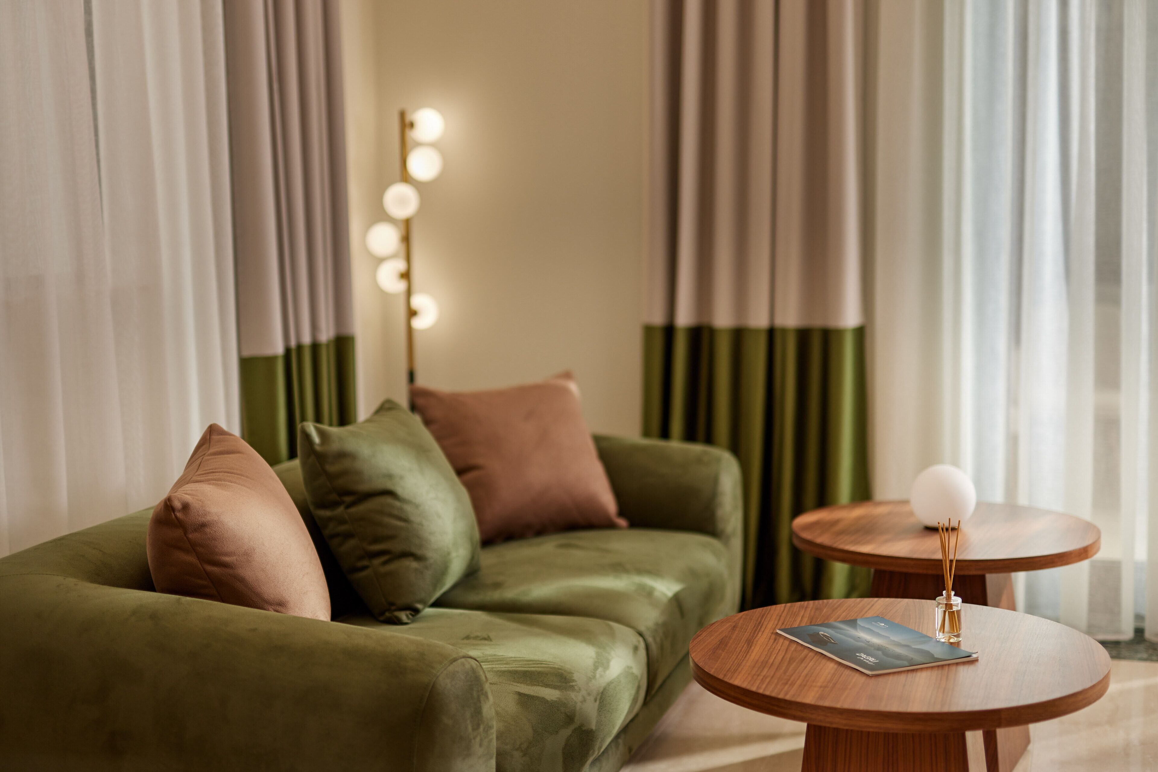 A green sofa with multiple cushions, a round wooden table with a magazine and a decorative item, and a backdrop of curtains and a light fixture.