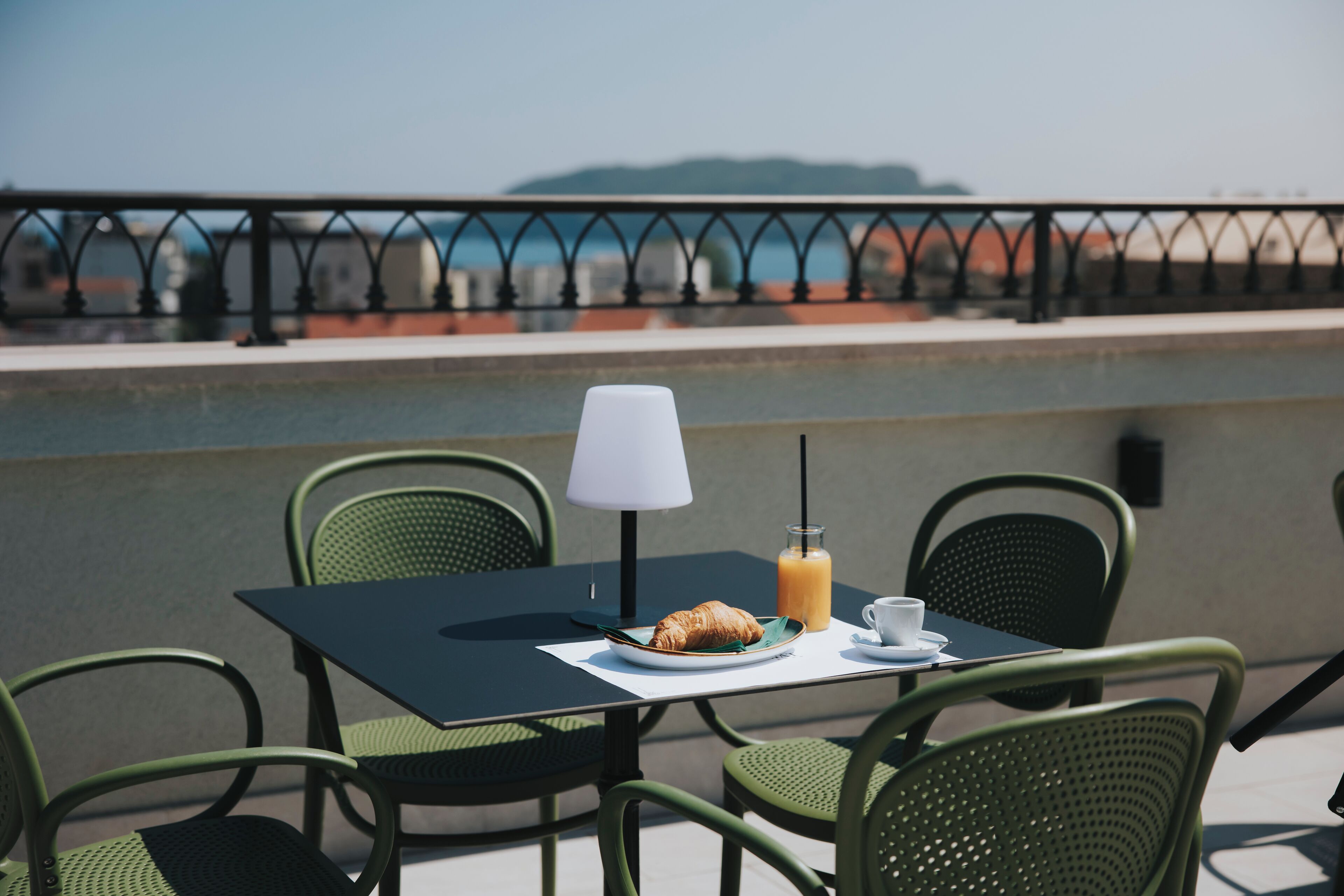 Una mesa con un croissant, un vaso de jugo de naranja y una taza blanca en una terraza con sillas verdes y un mantel negro.