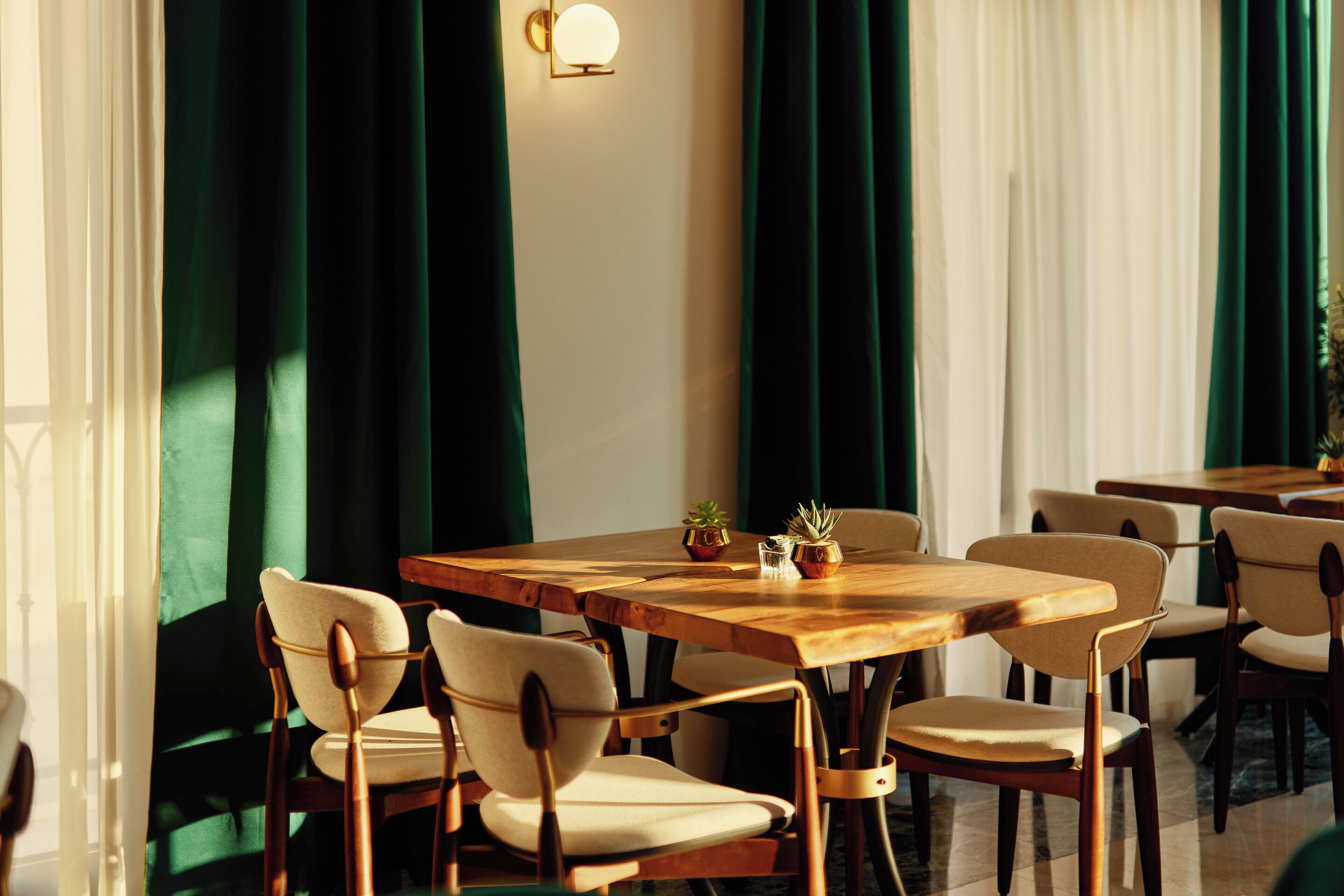 A dining area with wooden tables and chairs, a wall-mounted light fixture, and green curtains.
