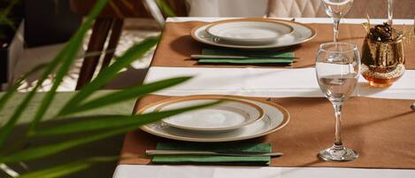A dining table set with plates, cutlery, and glasses, surrounded by chairs and a plant.