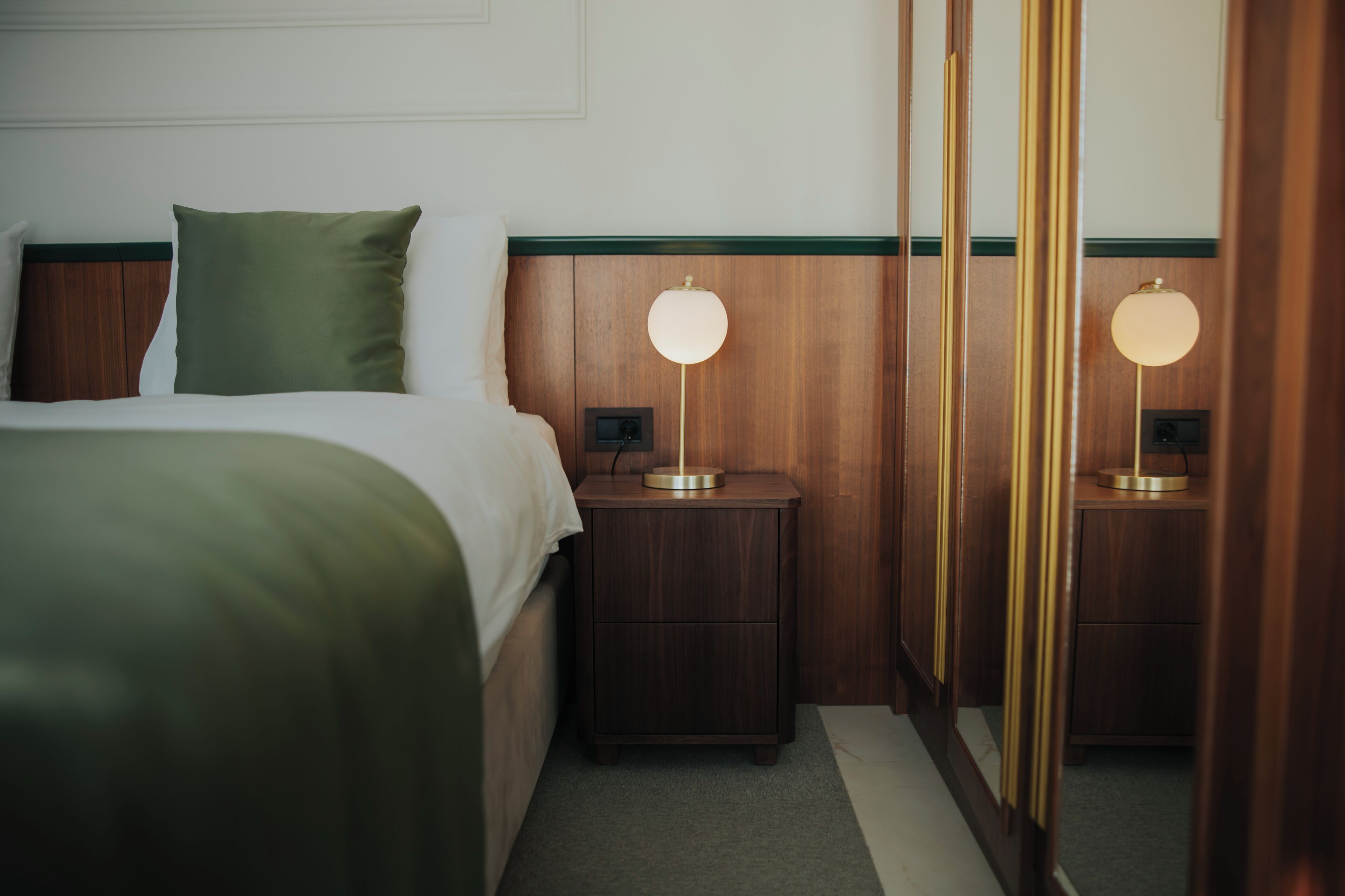 A neatly made bed with a green pillow, a bedside table with a lamp, and a mirror reflecting the room.