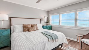 A bedroom with a large bed, a ceiling fan, a window with blinds, a nightstand, and a chair.