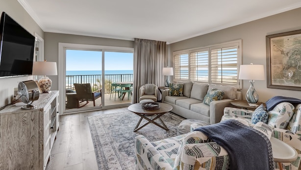 A spacious living room with a large flat-screen TV, a sofa set, a coffee table, and a view of the balcony.