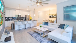Una sala de estar moderna con un sofá blanco, una mesa de centro y una cocina con gabinetes blancos y electrodomésticos de acero inoxidable.