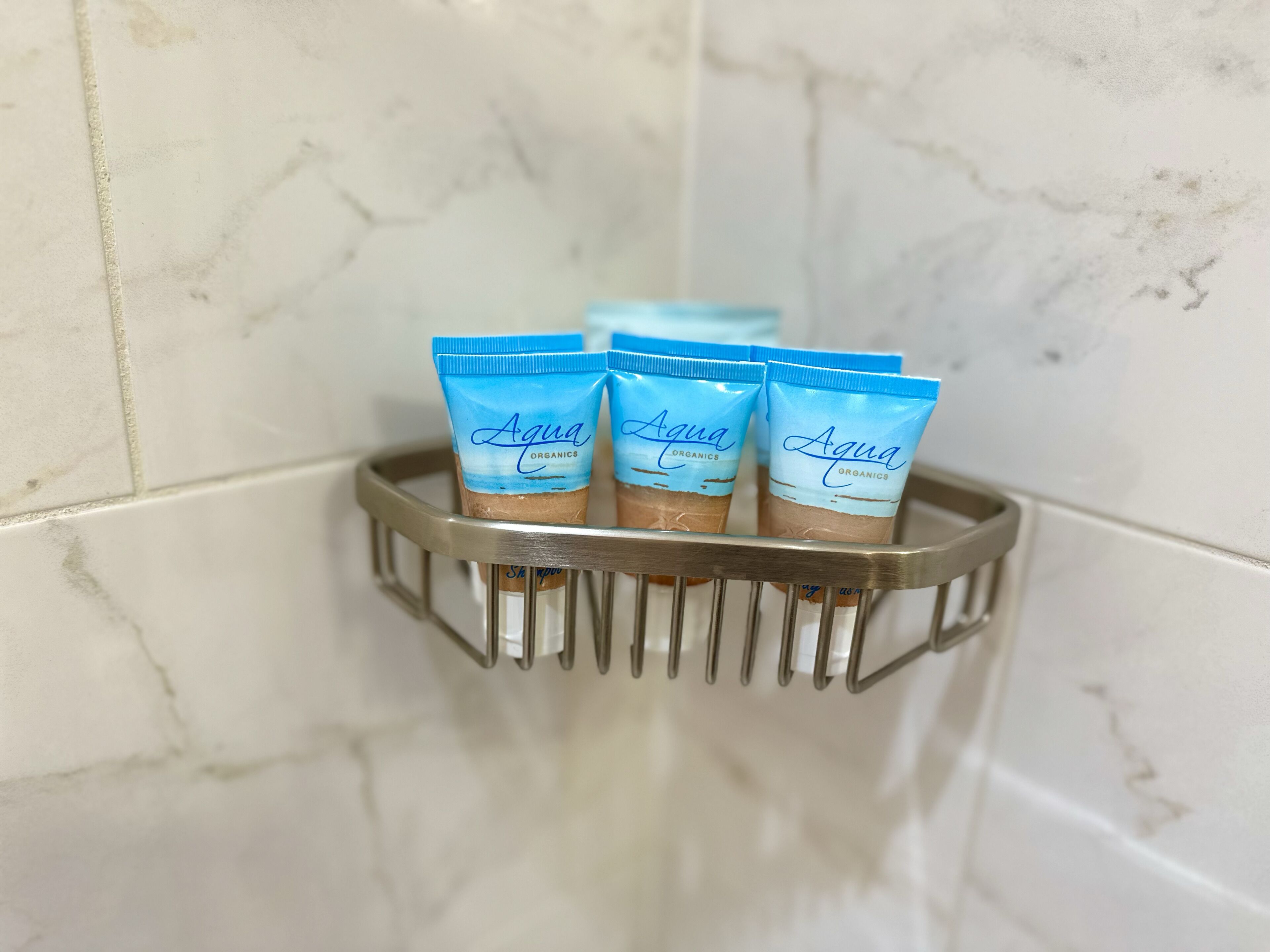 Una esquina de baño con una pared de mármol y un estante metálico que contiene cuatro tubos de champú Aqua Organics.