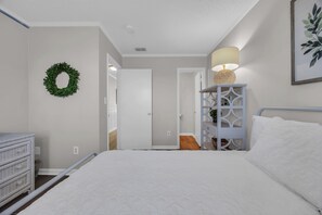 Une chambre à coucher comprenant un lit, une commode, une table de chevet avec une lampe, une étagère fixée au mur et une porte donnant sur une autre pièce.