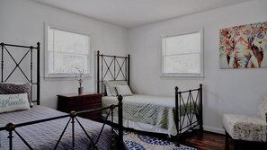 A bedroom with two single beds, a dresser, and a chair. There are windows with blinds, a painting on the wall, and a rug on the floor.