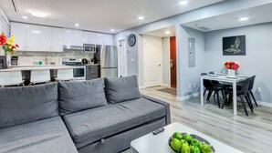 A modern apartment with a grey sofa, a dining table with chairs, a kitchen area with white cabinets and appliances, and a wall clock. - Bow 119 | Adorable Suite with Pool & Hot Tub (Canmore)