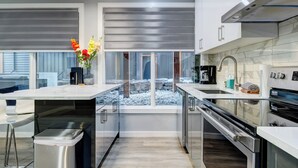 A modern kitchen with a central island, stainless steel appliances, and a view of a rocky outdoor area. - Bow 119 | Adorable Suite with Pool & Hot Tub (Canmore)