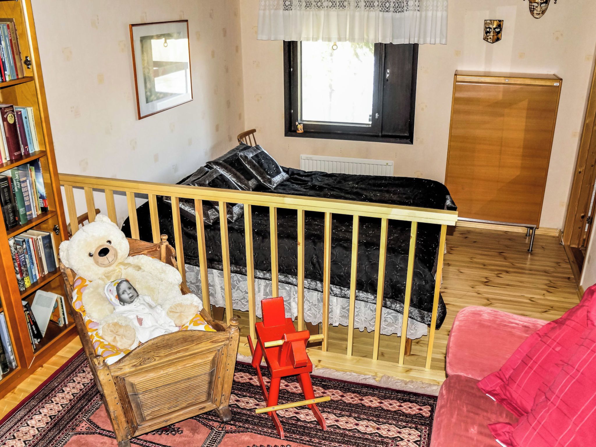 Habitación infantil con una cuna, una estantería, una silla roja, un sofá rosa y una ventana con vistas.
