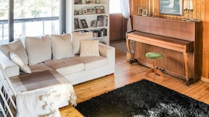 Ein gemütliches Wohnzimmer mit einem weißen Sofa, einem Holzpiano und einer Bücherwand voller Fotos und Bücher.