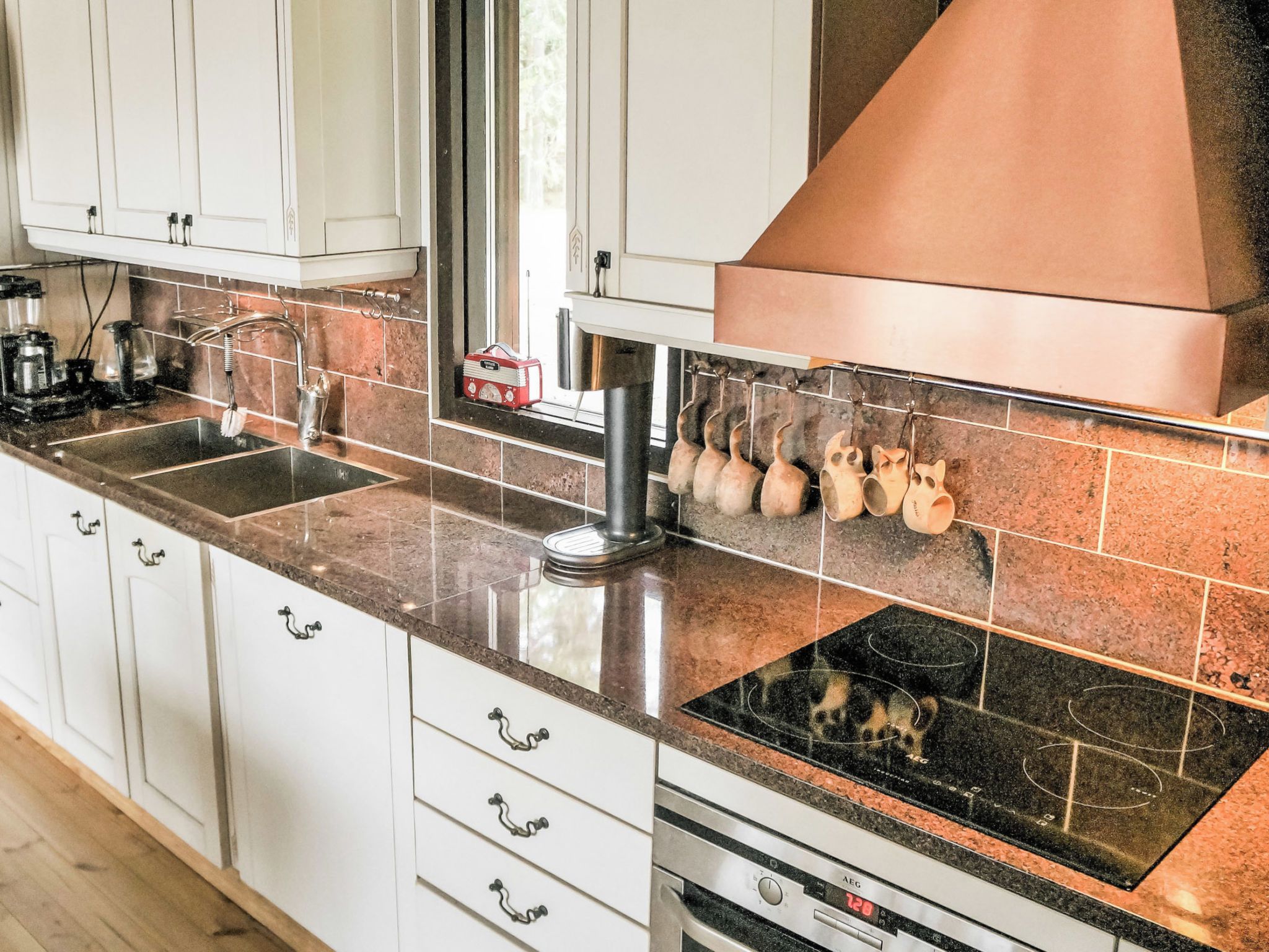 A kitchen with white cabinets, a stainless steel range hood, a black induction cooktop, and a window above the sink.