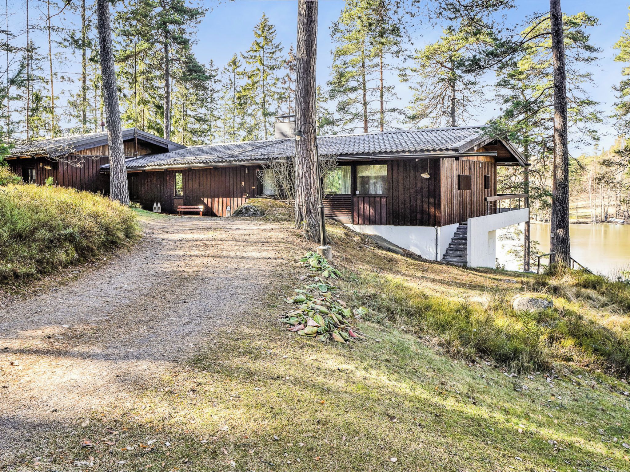 Un chalet rustique, accessible par un chemin de gravier, entouré de grands arbres et bordé par une étendue d’eau.
