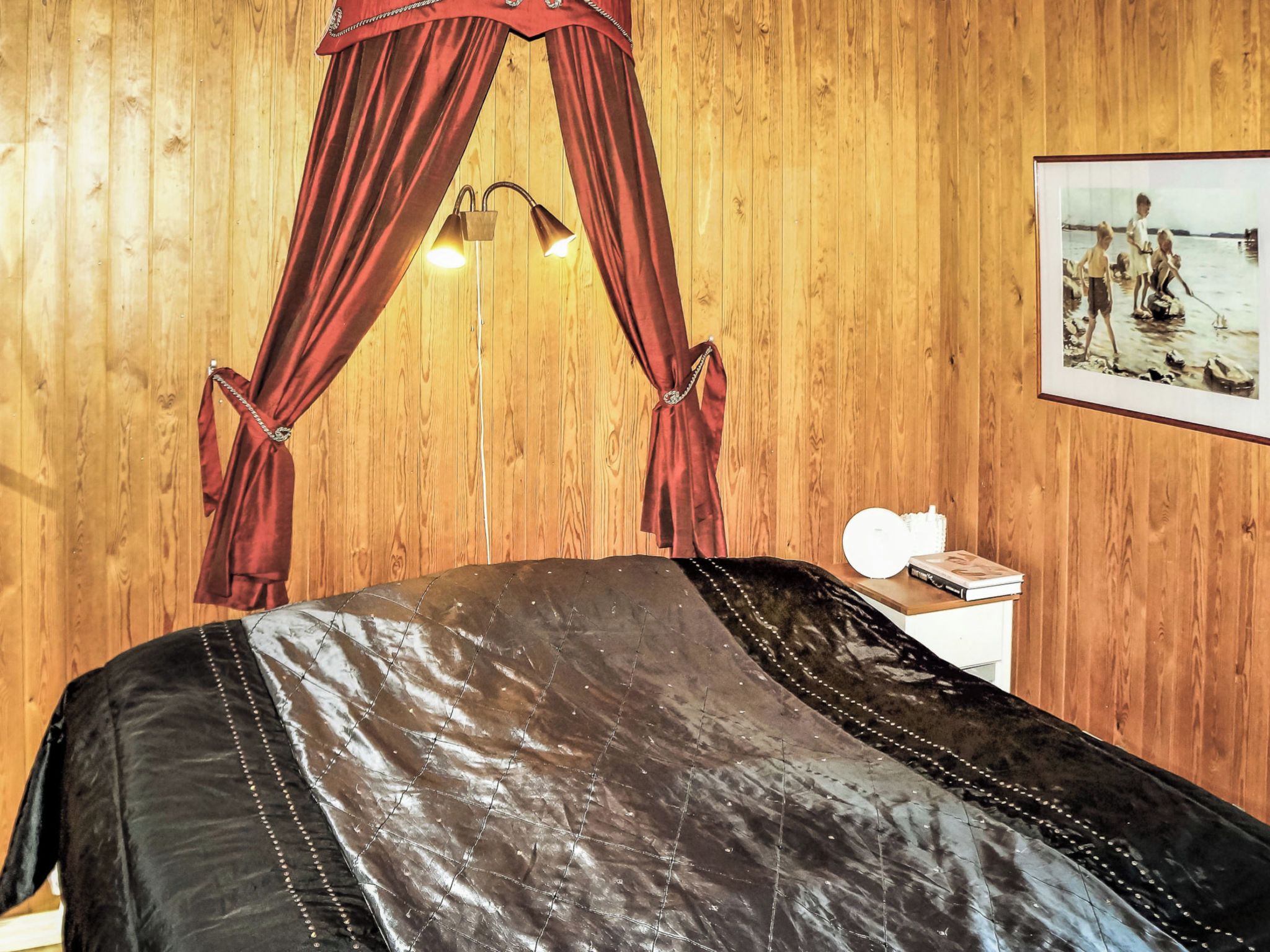 A room with a bed, a wooden wall, a framed picture, and a bedside table with a lamp.
