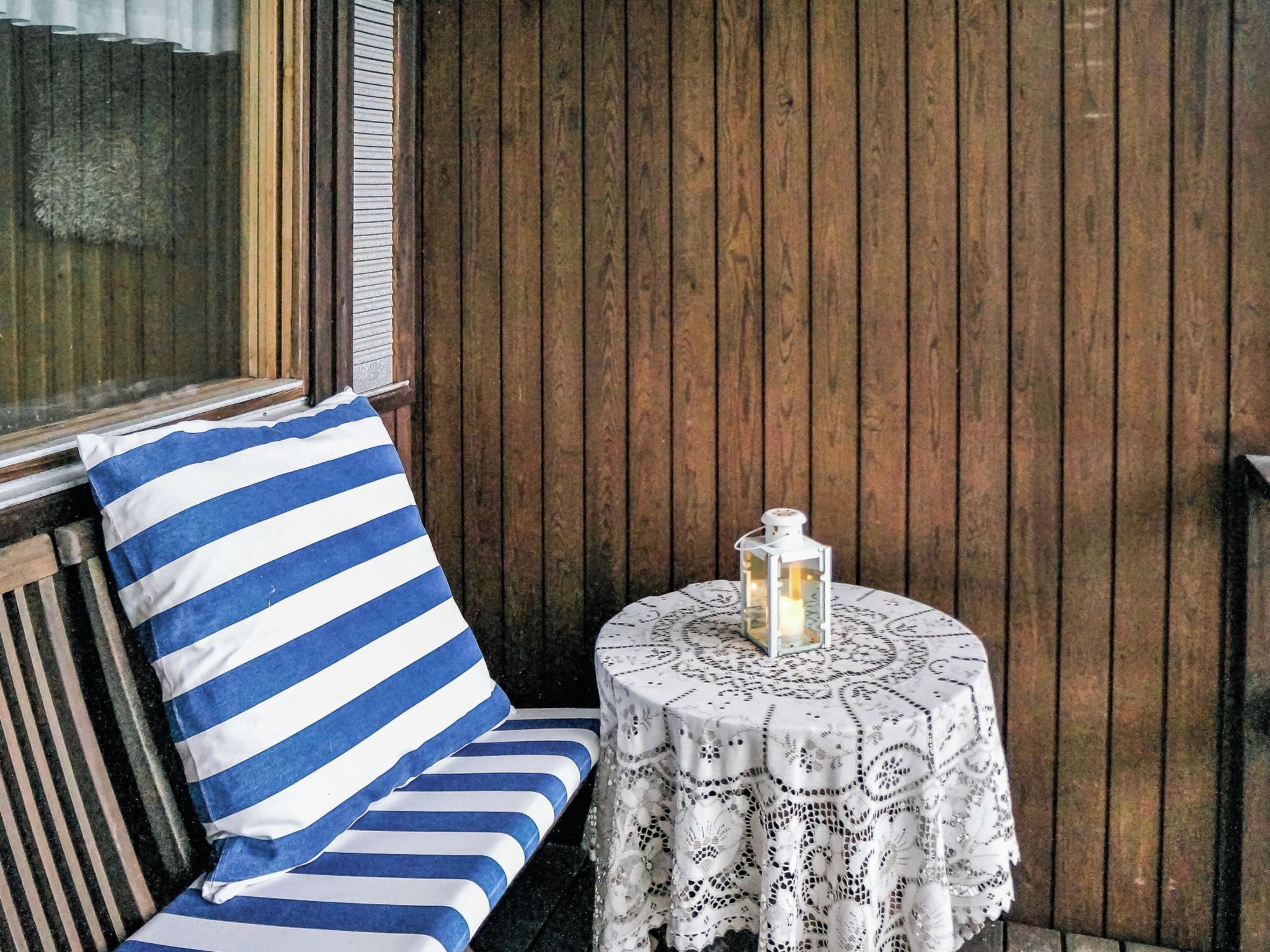 A wooden bench with a blue and white striped cushion, a small round table with a lace tablecloth, and a white lantern on the table.