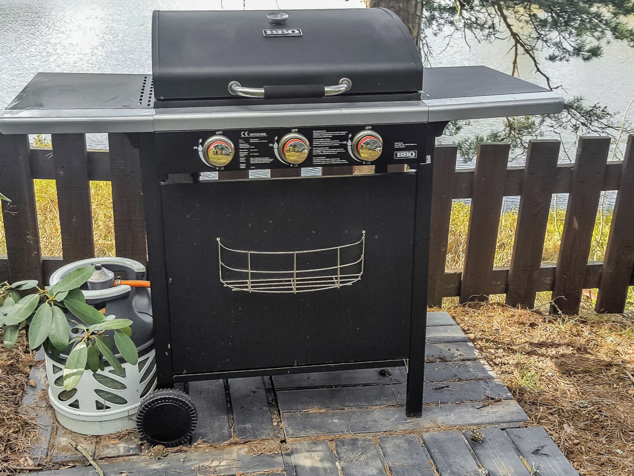 A portable grill with control knobs and a metal grate design, placed on a paved surface with a wooden fence in the background.