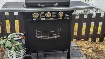 A portable grill with control knobs and a metal grate design, placed on a paved surface with a wooden fence in the background.