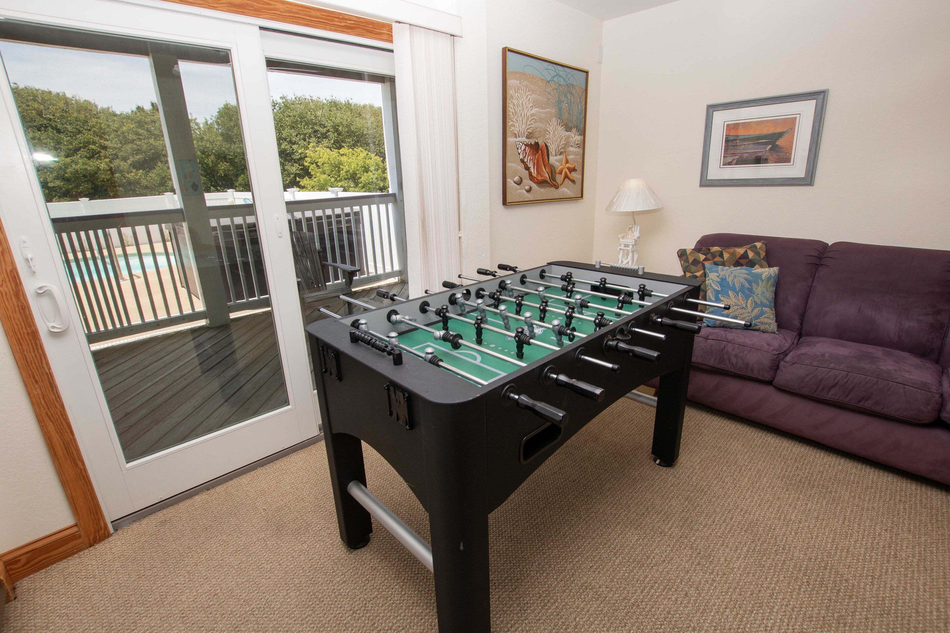 A foosball table in a room with a purple sofa, a painting, and a view of a balcony with a pool.