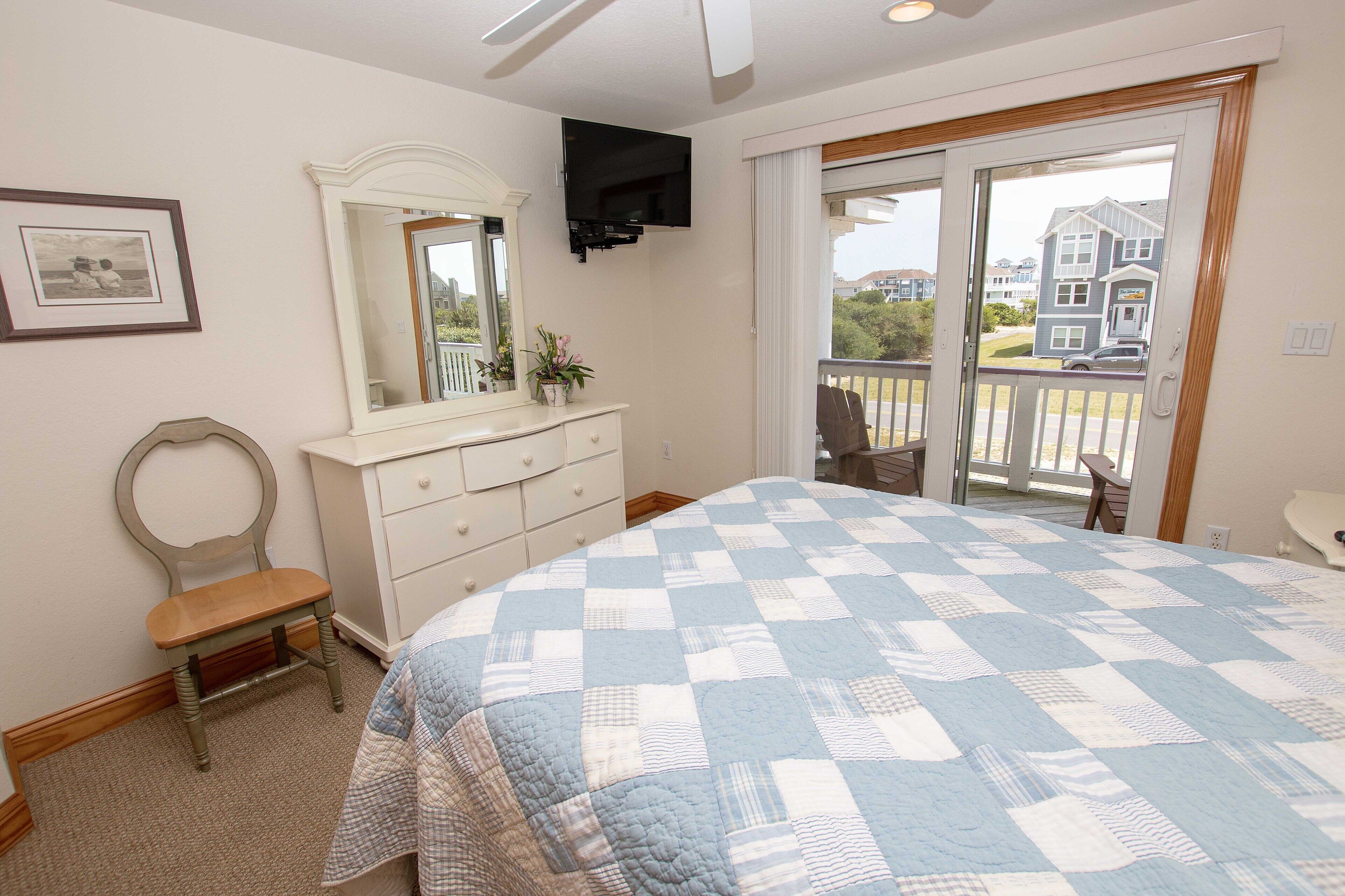 A bedroom with a large bed, a dresser, a chair, a TV mounted on the wall, and a balcony with a view of houses.