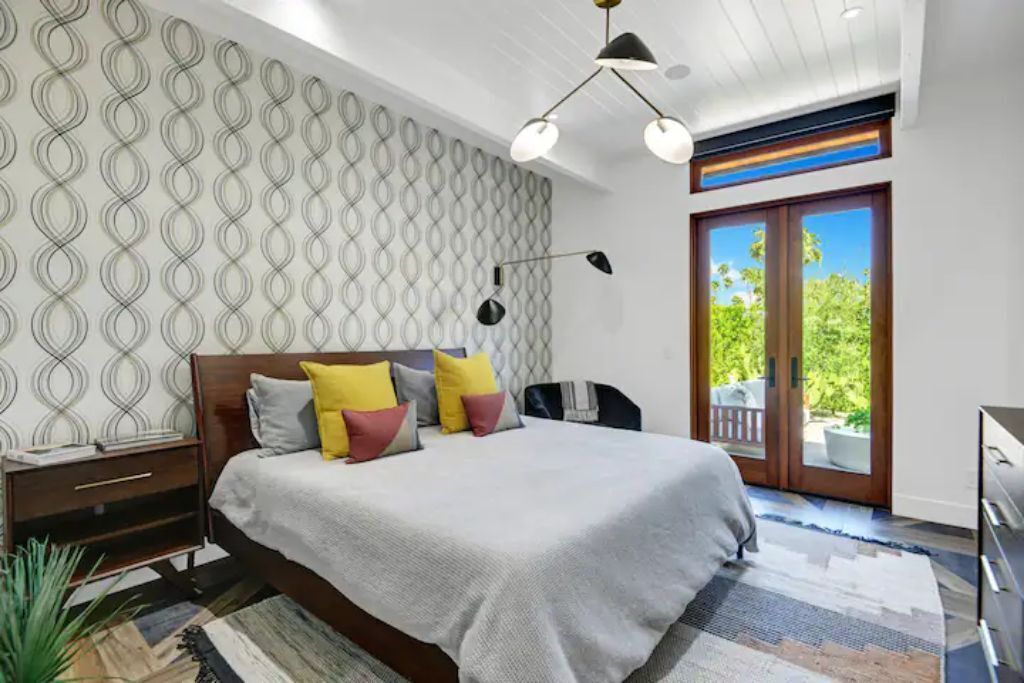 A modern bedroom with a large bed, a patterned wallpaper, a ceiling light, and a view of the outdoors through a glass door.