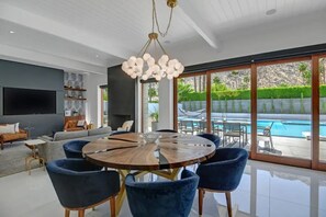 Ein modernes Wohnzimmer mit einem runden Holztisch, blauen Stühlen, einem großen Fernseher und Blick auf einen Pool.