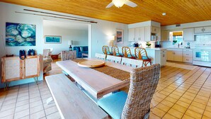 Un amplio espacio habitable con techo de madera, una mesa de comedor con sillas de mimbre, una cocina al fondo y un cuadro grande en la pared.