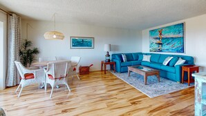 Una sala de estar con un sof á azul, sillas de mimbre blanco, una mesa de centro de madera, una lámpara y un cuadro de un surfista.
