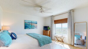 Un dormitorio con cama, ventilador de techo, ventana con cortinas, mesita de noche y espejo.