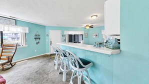 Una cocina con barra, taburetes blancos, ventilador de techo y vistas al exterior.
