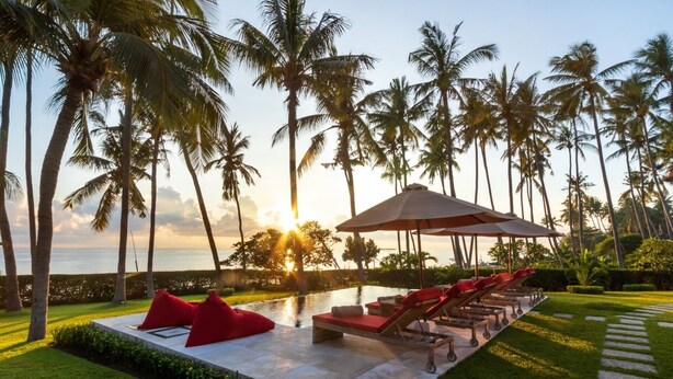 A poolside area with lounge chairs and red cushions, surrounded by palm trees and a view of the ocean at sunset. - Renovated 3BR Villa W/ Pool - Perfect for Couples , Bali Villa 2237 (Bali)