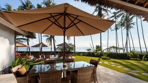 An outdoor dining area with a glass table and wicker chairs, surrounded by palm trees and a view of the ocean.