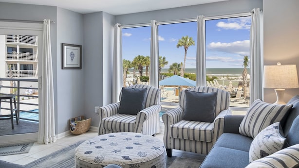 A modern living room with striped furniture, large windows with curtains, a view of a pool and palm trees, and a beach in the background. - Oceanfront at Villamare! Pool and Ocean Views. (Hilton Head Island)