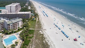 A beachfront hotel with a long sandy beach, a swimming pool, and a large building with multiple balconies. - Oceanfront at Villamare! Pool and Ocean Views. (Hilton Head Island)