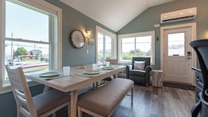A dining area with a wooden table, chairs, and a bench. There are windows with white frames and a view of the outside.